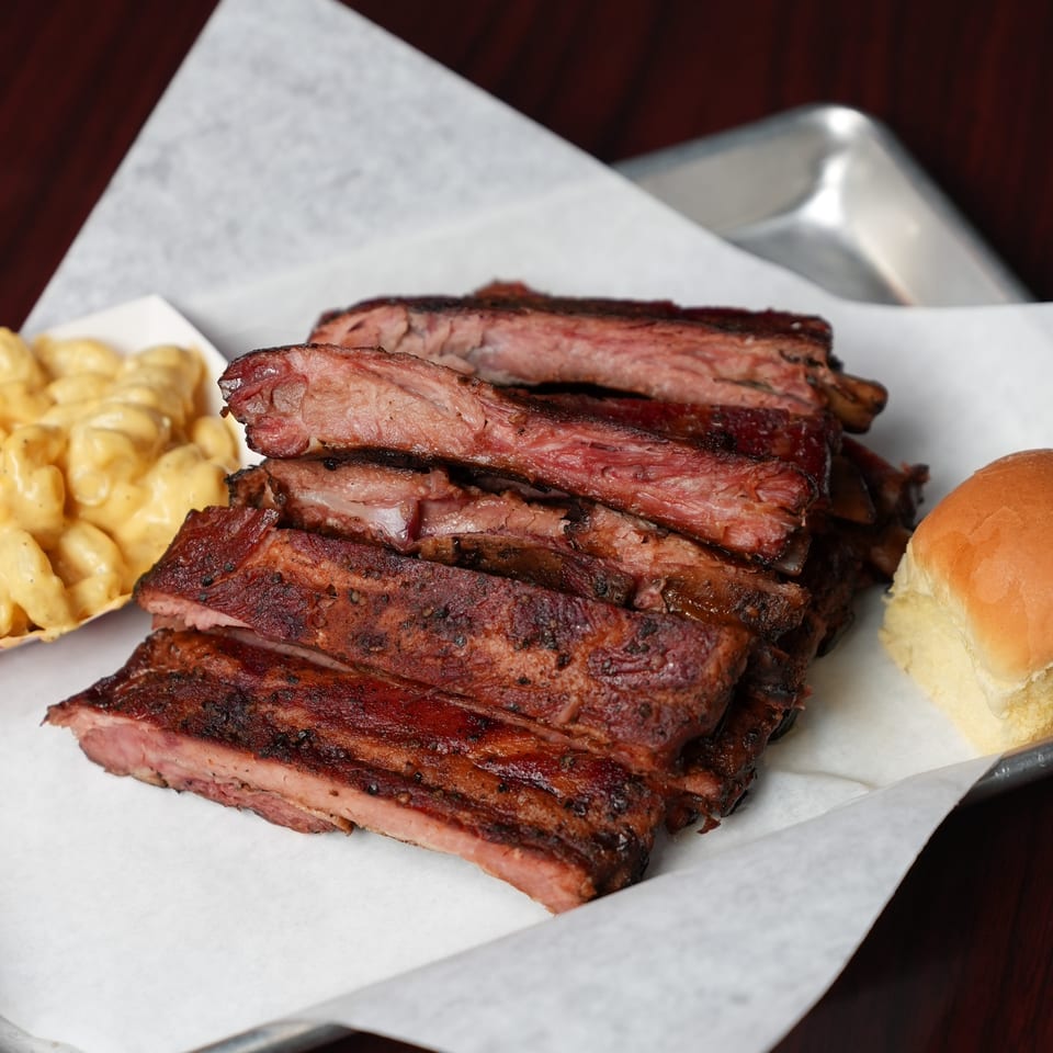 Stack of smoked barbecue ribs served with creamy macaroni and cheese and a soft bread roll on a paper-lined tray.
