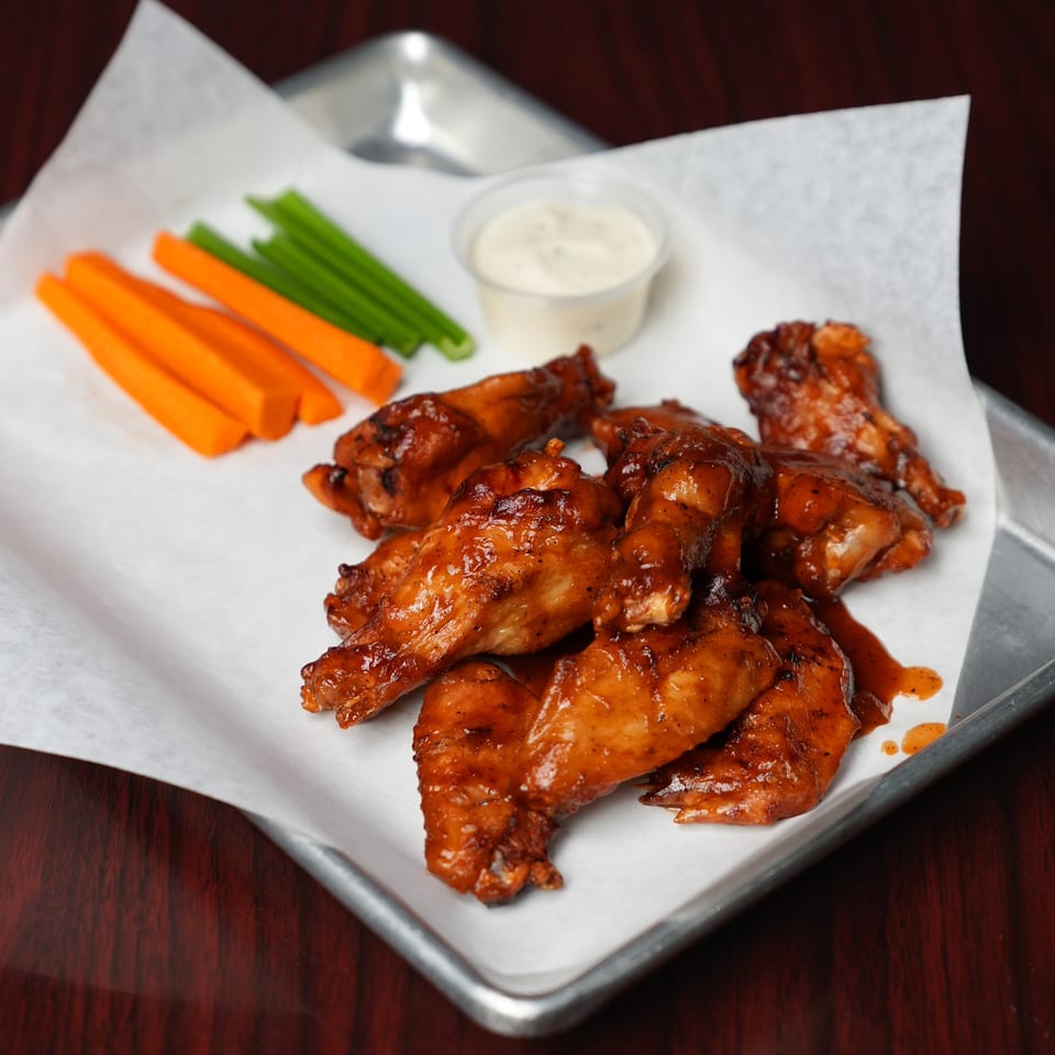 Glazed chicken wings served with carrot and celery sticks and a cup of creamy dipping sauce on a metal tray.