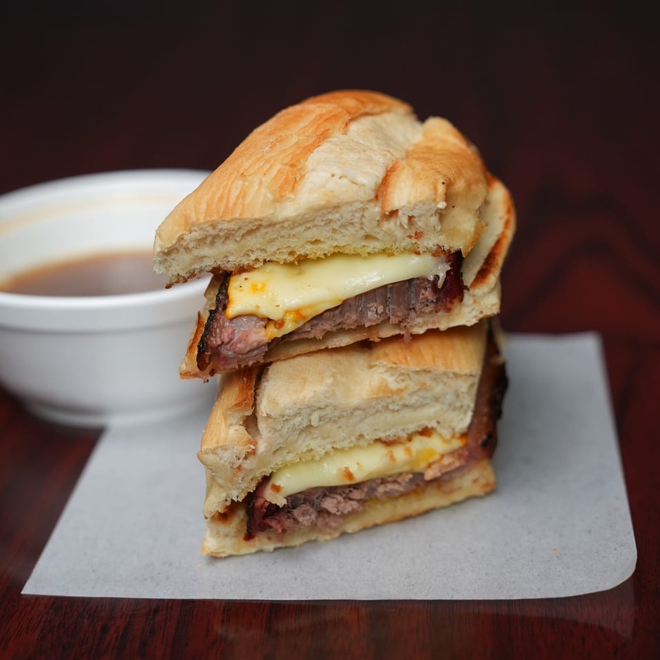 Stacked roast beef sandwiches with melted cheese on toasted bread, served with dipping sauce on a white plate.