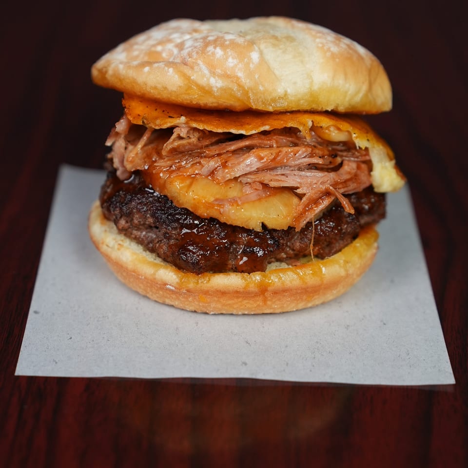 Gourmet burger on a brioche bun layered with a beef patty, pulled meat, melted cheese, and crispy onion rings.