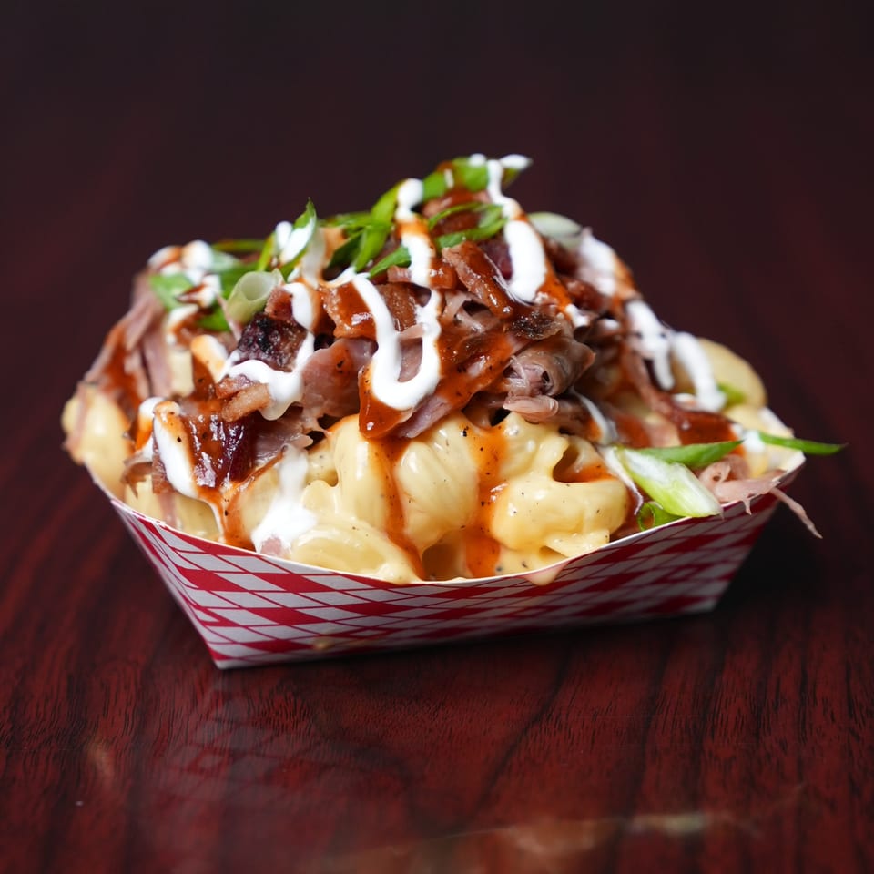 Loaded fries topped with sliced meat, melted cheese, white and red sauces, and green onions in a paper tray.