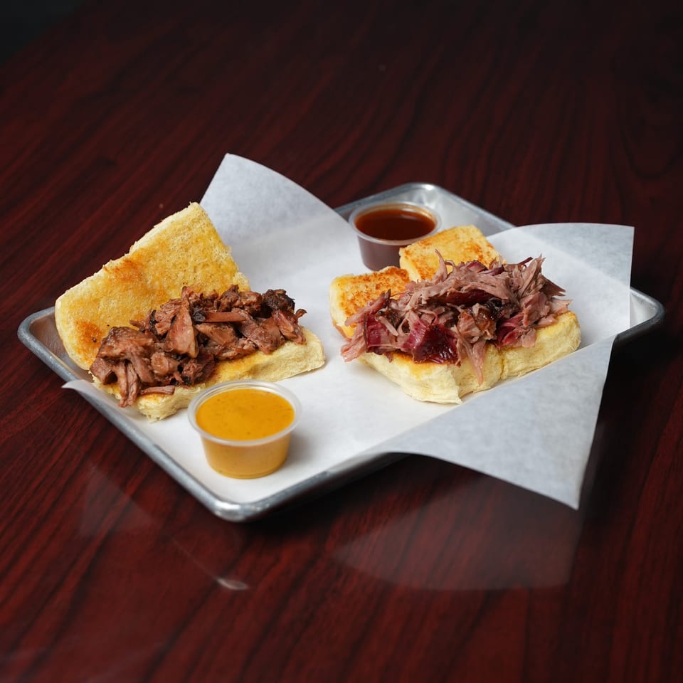 Two toasted sandwiches filled with shredded meat sit on a lined metal tray with dipping sauces on a wooden table.